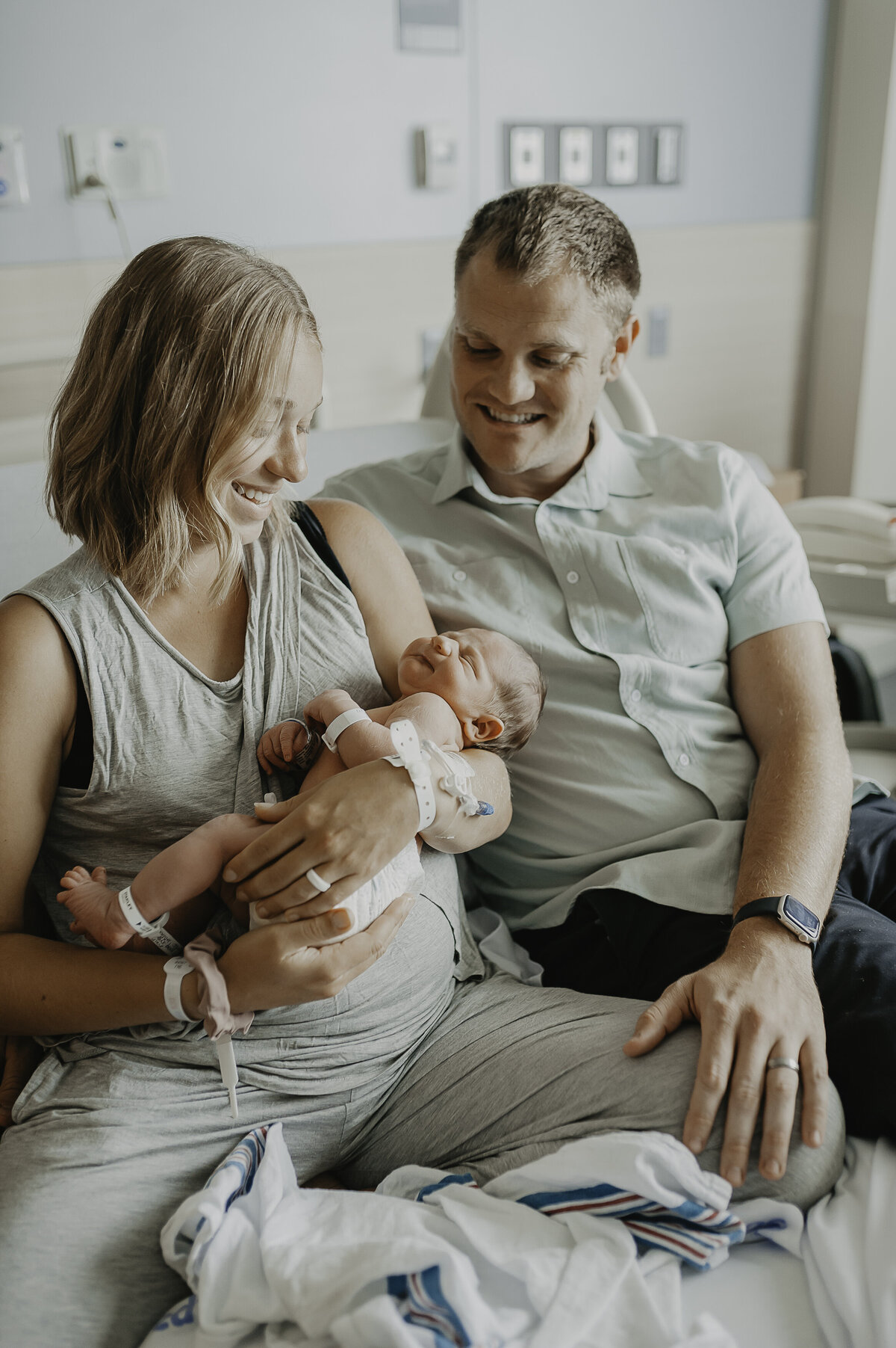 Verona B Photography captures parents gazing at their newborn baby while sitting on a hospital bed. Mom still has her IV in but looks amazing. Mom and dad look totally captivated with the newest addition to their family. There is something awe-inspiring about these Fresh 48 sessions as they capture the newness of bringing another little life into the world – Akron General Hospital session.