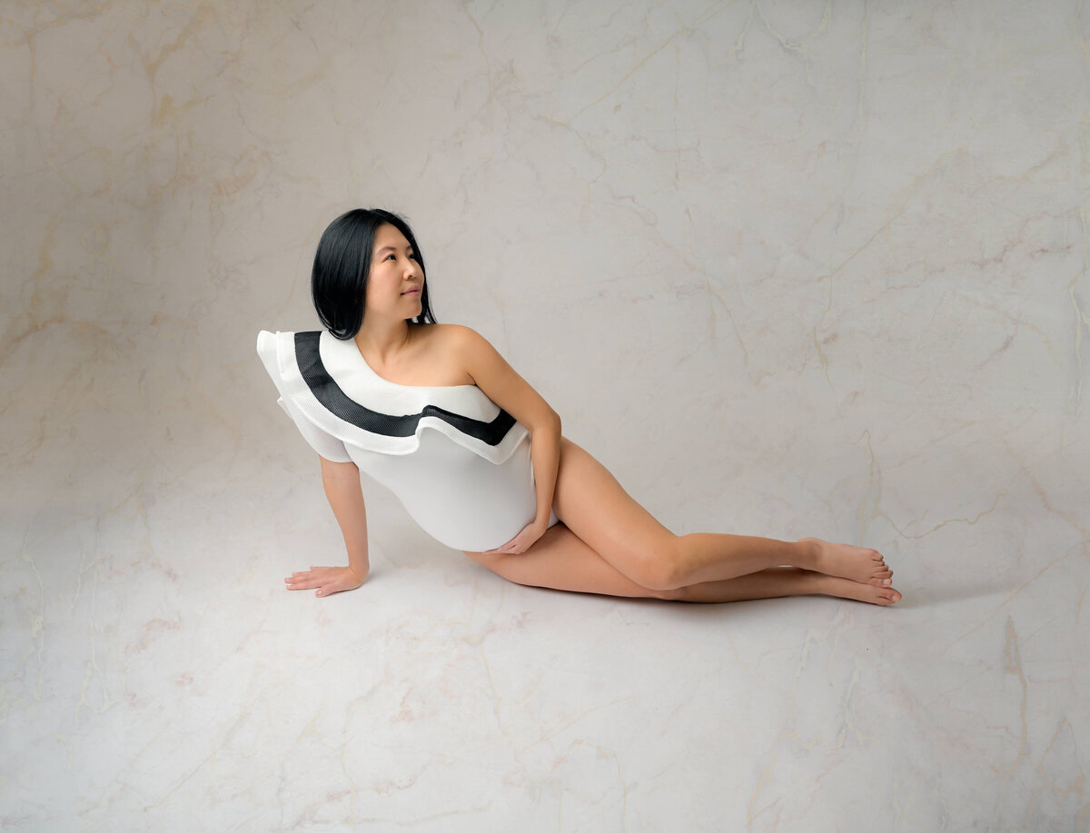 Maternity studio session where mom-to-be is sitting on the floor on her side and looking over her shoulder.  She's wearing an elegant white bodysuit. Image captured by Beth Benecki