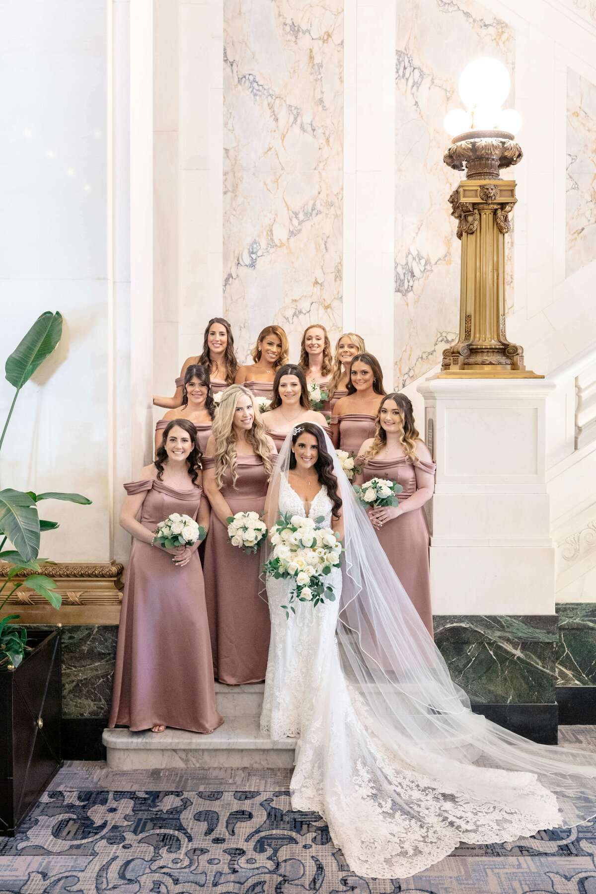 A bride in a lace gown and long veil stands with eight bridesmaids in mauve dresses on a grand staircase. They all hold white and green bouquets. The background features marble walls and an ornate lamp.