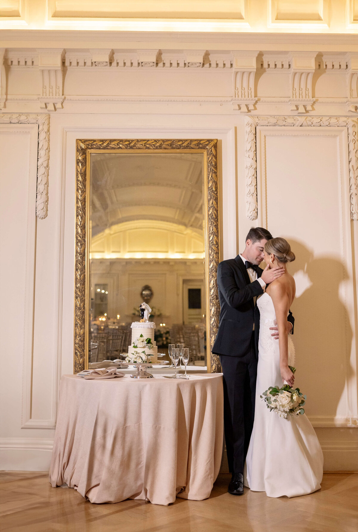 A bride and groom embrace in front of a large mirror in an elegant room. A table with a two-tiered wedding cake and flowers is nearby. The bride holds a bouquet, and both are dressed in formal attire.