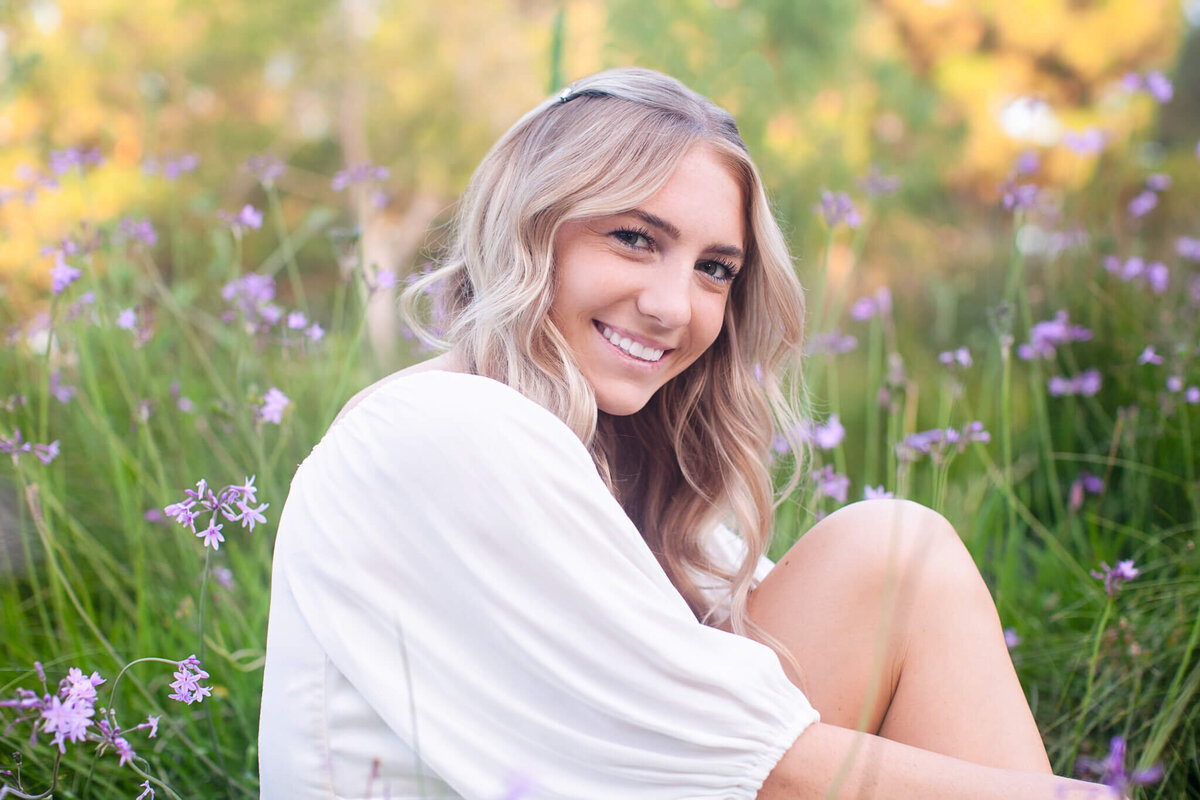 A woman with long, wavy blonde hair sits in a field of purple flowers, wearing a white blouse. She is smiling and surrounded by greenery, with soft, warm lighting suggestive of a late afternoon or early evening.
