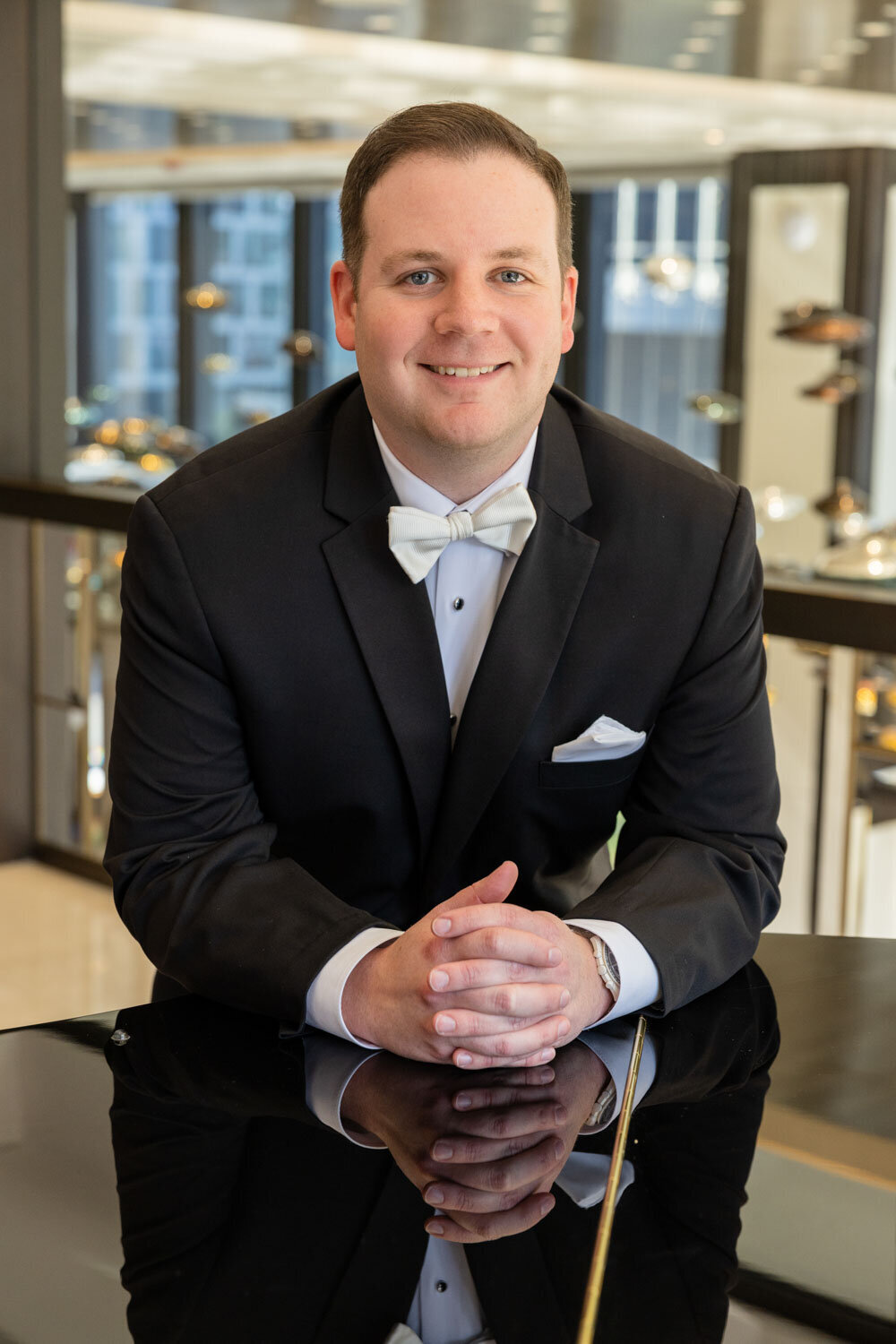 A man in a tuxedo and bow tie is sitting at a table with his hands folded