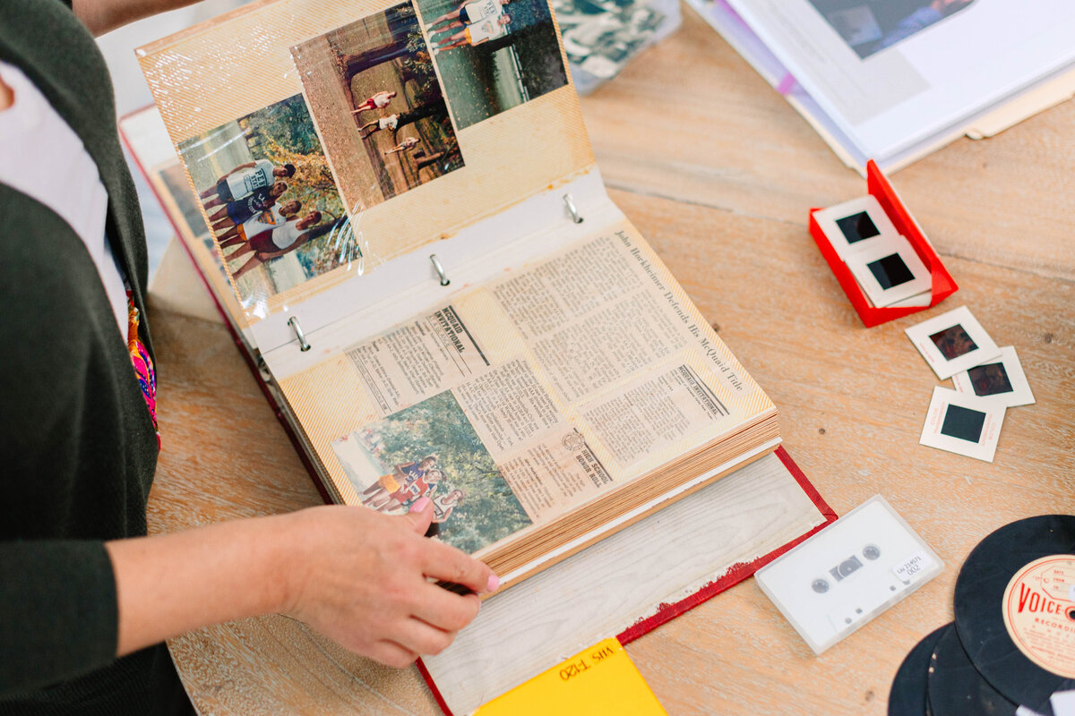 A person flips through a vintage scrapbook filled with old photographs and newspaper clippings on a wooden table. Surrounding the book are nostalgic items, including film slides, a cassette tape, and a vinyl record.