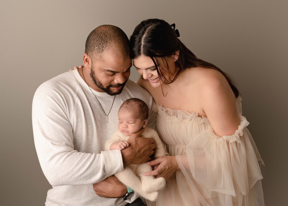 Mom and dad pose for an image at their newborn's photoshoot. Dad is holding the baby and mom is standing slightly behind him. Both are looking down at the baby and smiling. Captured by best Corona newborn photographer Bonny Lynn Photography.