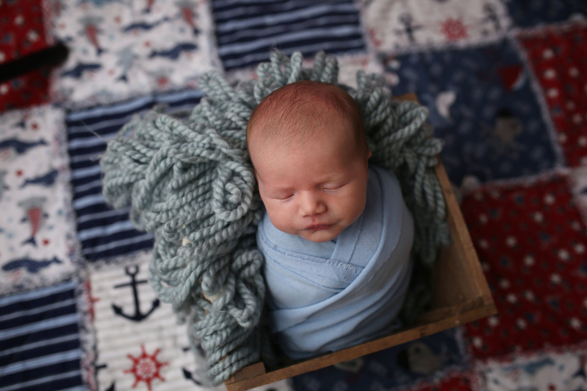 newborn baby boy wrapped tightly in blue cloth posed in wooden box prop