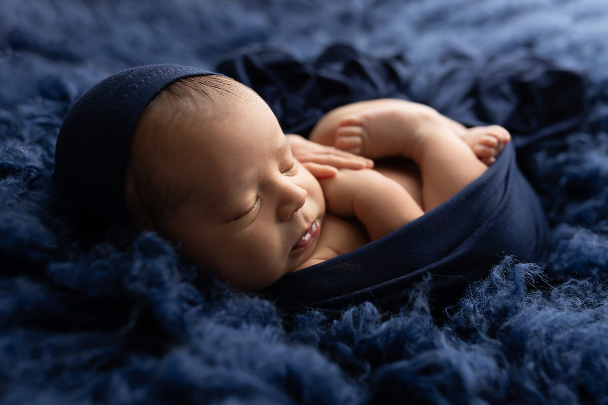 Sweet newborn baby peacefully sleeping wrapped in navy blue fabric on soft blue textures by Dallas newborn photographer.