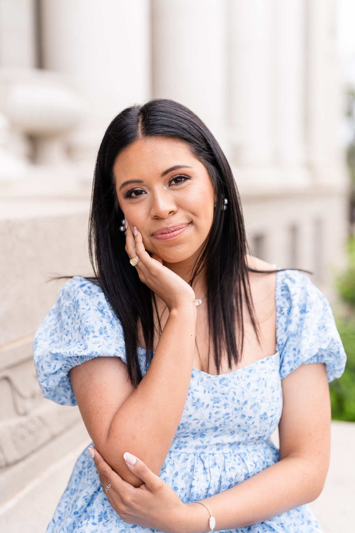 Texas A&M senior girl holding elbow and hand on cheek while wearing blue dress and sitting in front of Administration Building