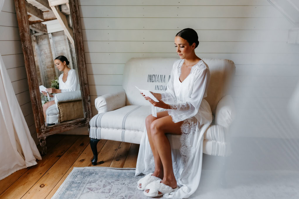 Bride sits on a couch in a white robe and fuzzy slippers and reads her vows in front of her Minneapolis wedding photographer