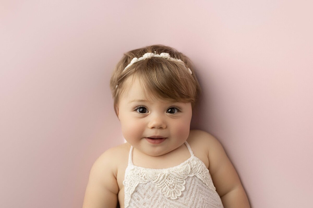 Aerial image of one-year-old baby in lace-trimmed crocheted top and coordinating headband smiling at the camera on pink plush backdrop.