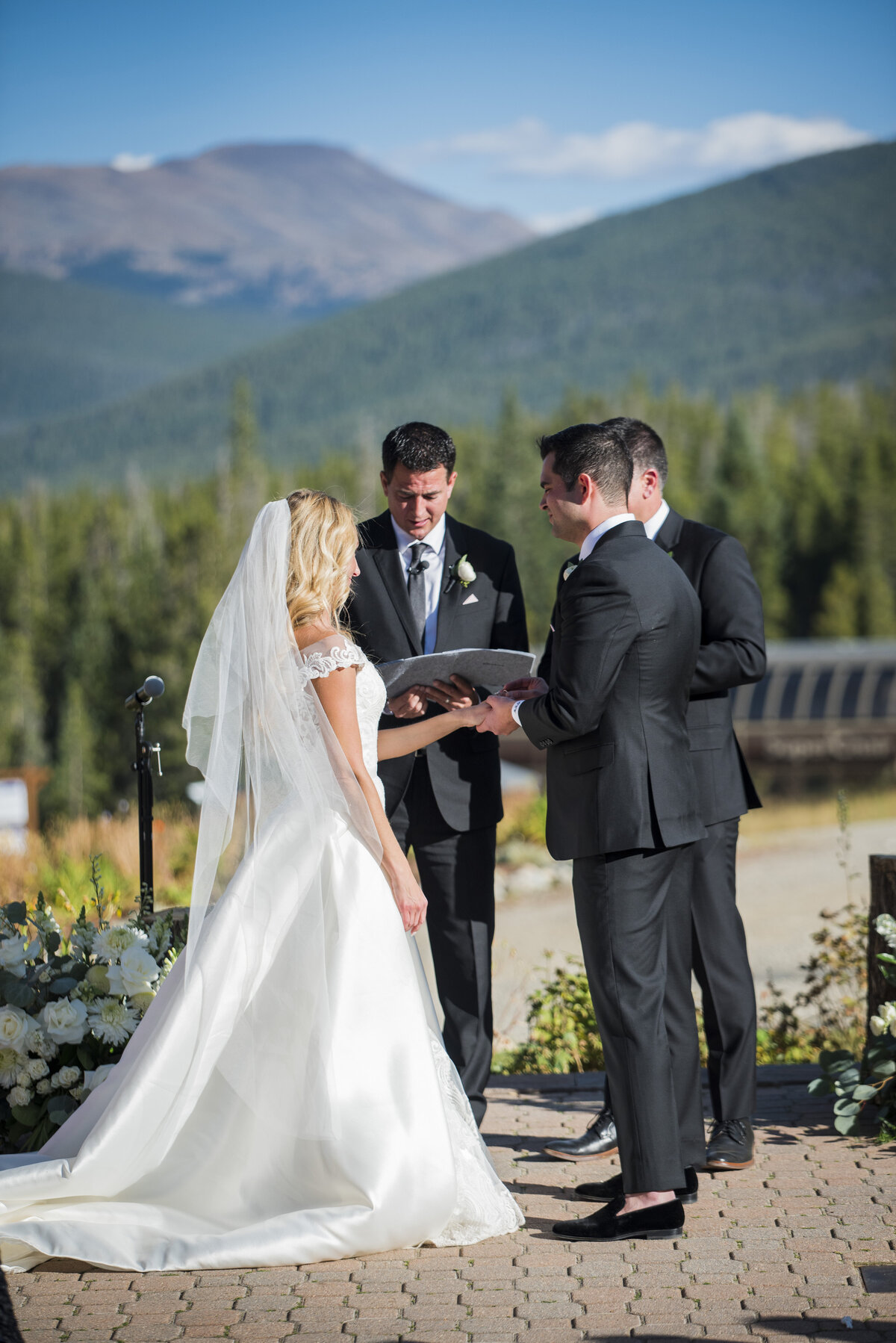 A groom holds his bride's hand during the outdoor wedding ceremony in Denver, Colorado with a mountain landscape in the background.