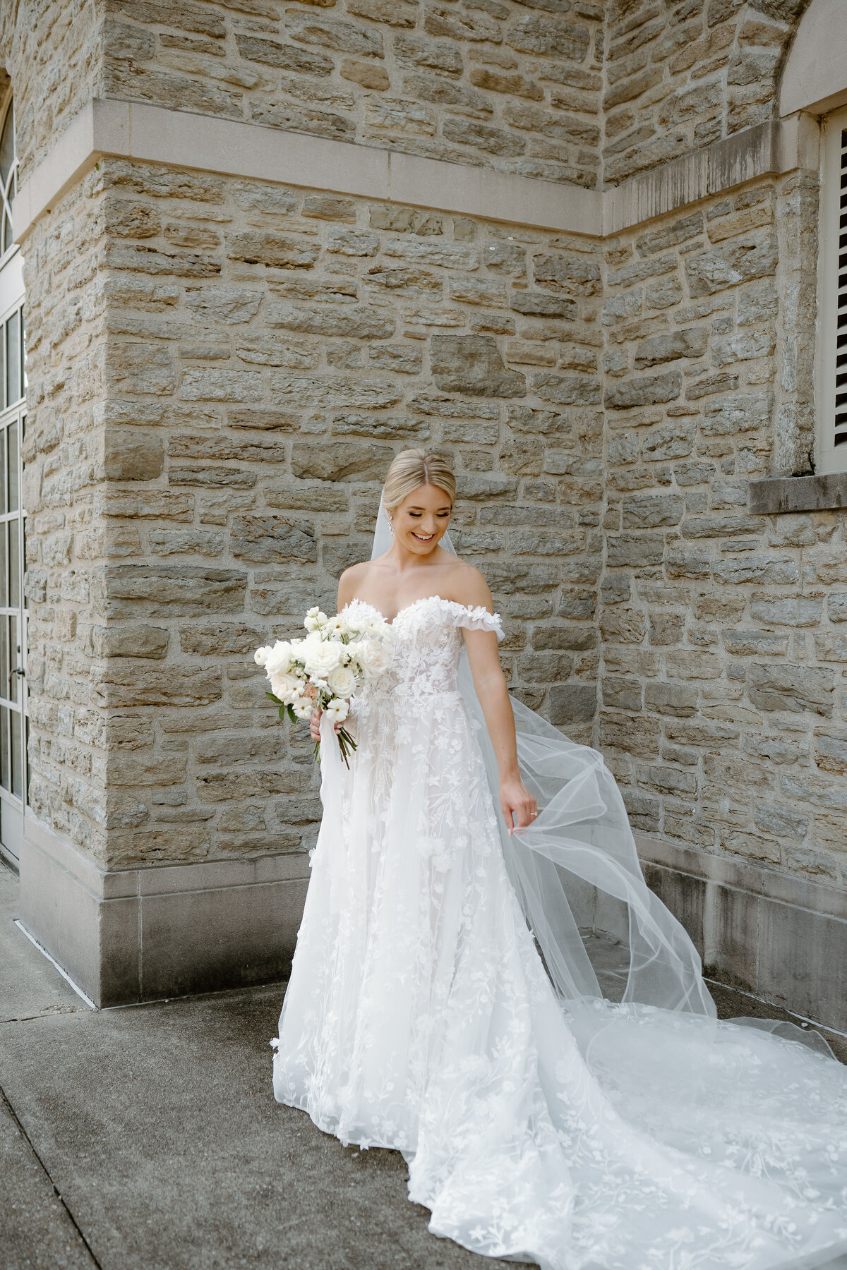 Bride in floral lace gown with bouquet, posing outside stone building.