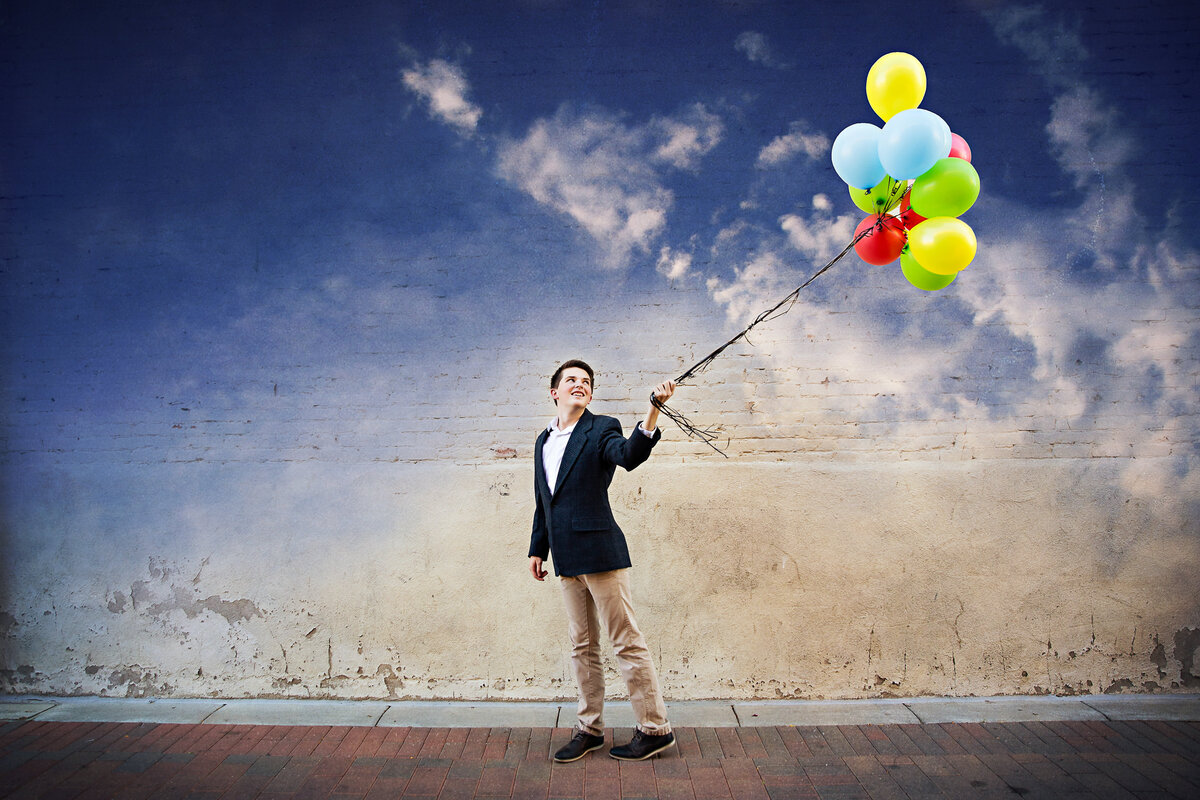 A high school guy in a suit jacket with a bunch of balloons in an urban setting