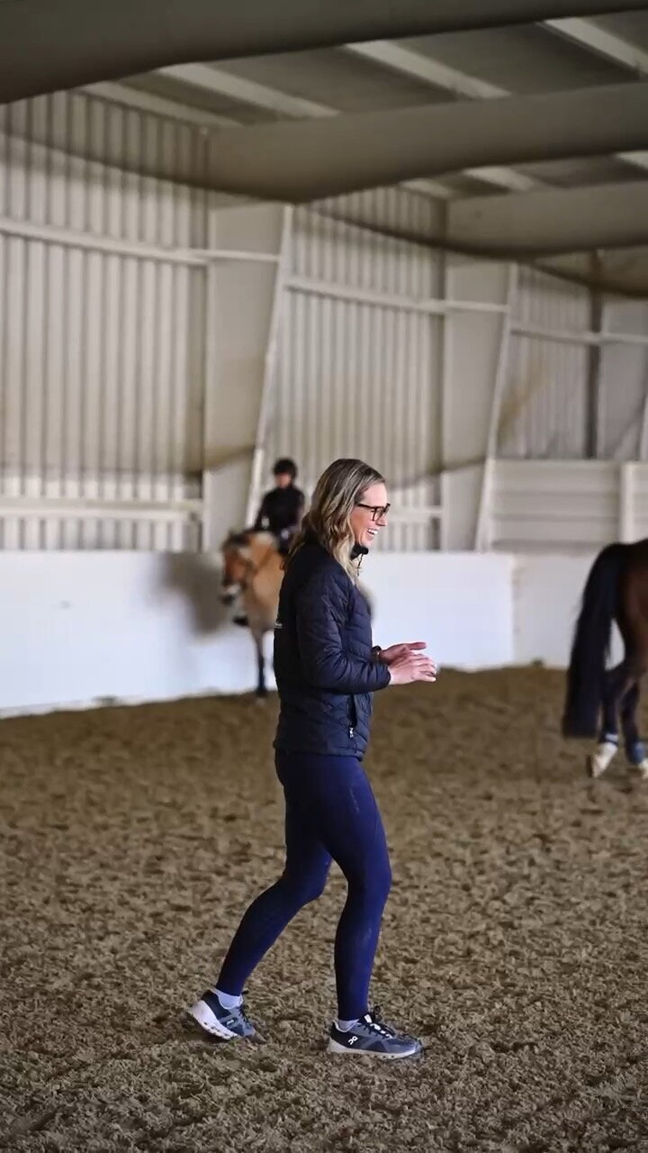 A riding instructor giving a group lesson to adult-amateur instructors, she is smiling while horses and riders pass by in the background