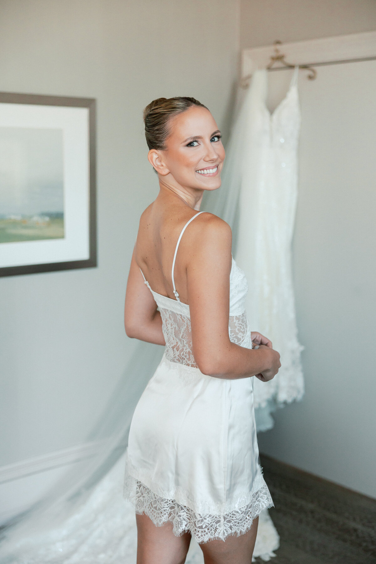 Bride smiling in a white lace bridal slip with her gown hanging in the background, shot in Philadelphia.