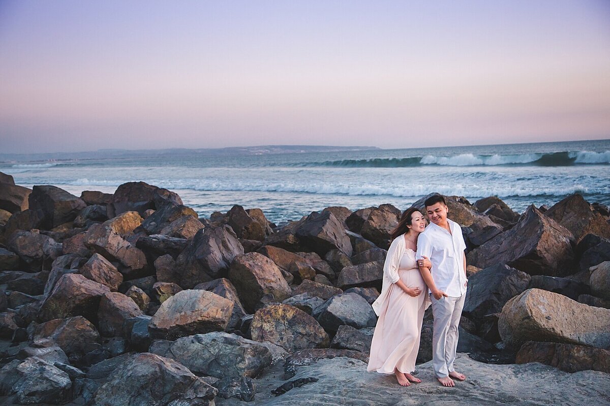 photo of a dad and pregnant mom, they are dressing in light colors and standing together on the sand with large rocks and the ocean behind them.