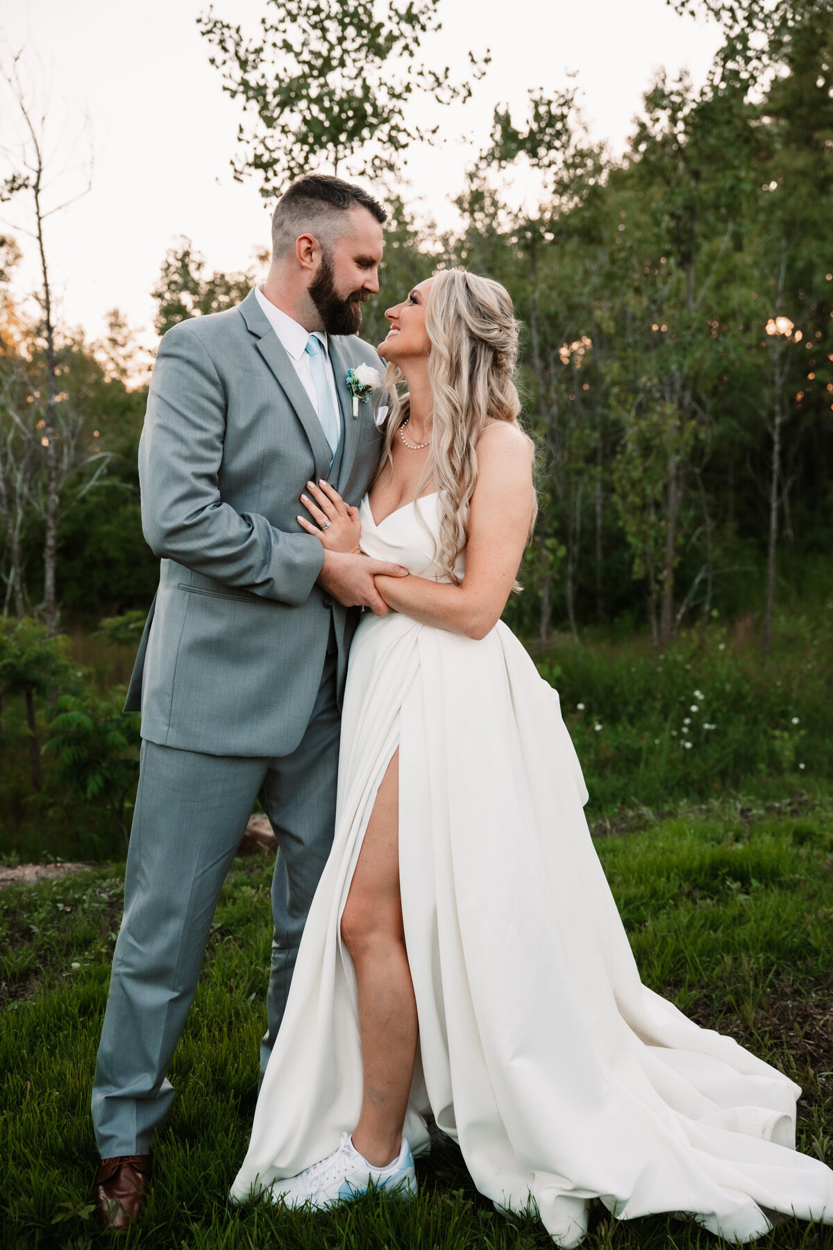 A bride and groom snuggle while standing in a lawn at sunset on edge of forest