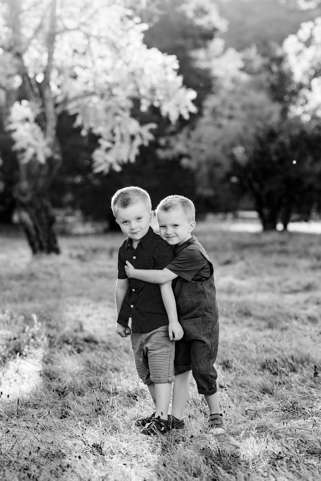 Twin boys stand in a field, one twin hugging the other, both looking at the camera