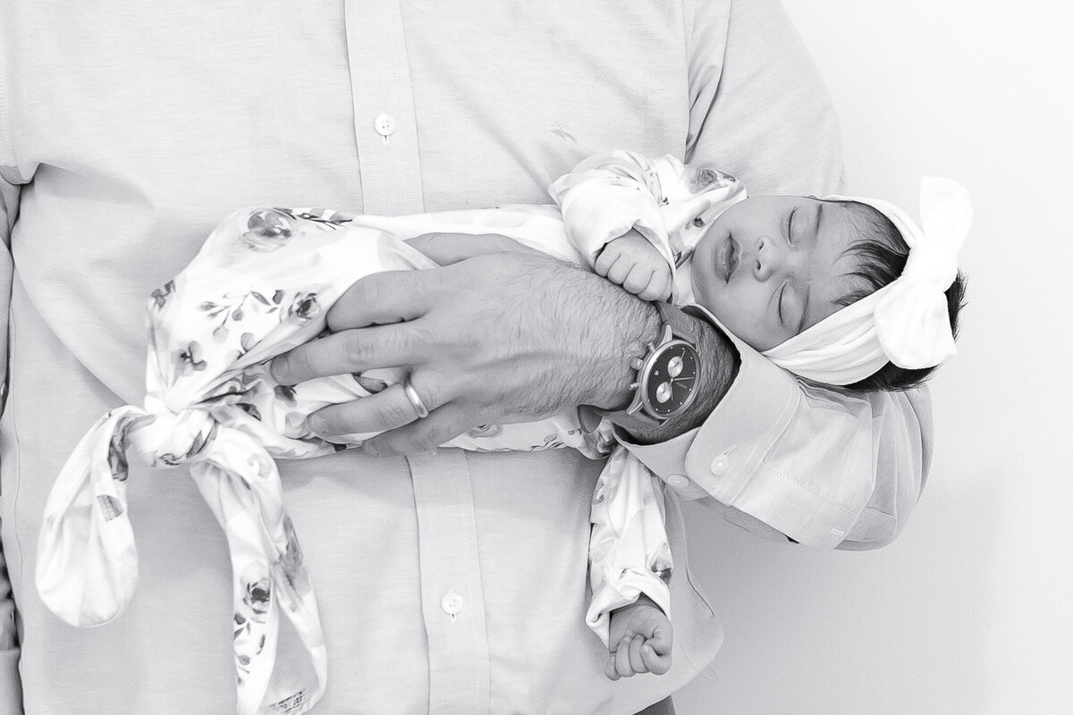 Black and white newborn photography of a sleeping baby girl wrapped in a floral swaddle, cradled in her father's arms. Captured by Birmingham newborn photographer Elle Lovelace, this timeless image highlights the delicate beauty of a baby’s early days