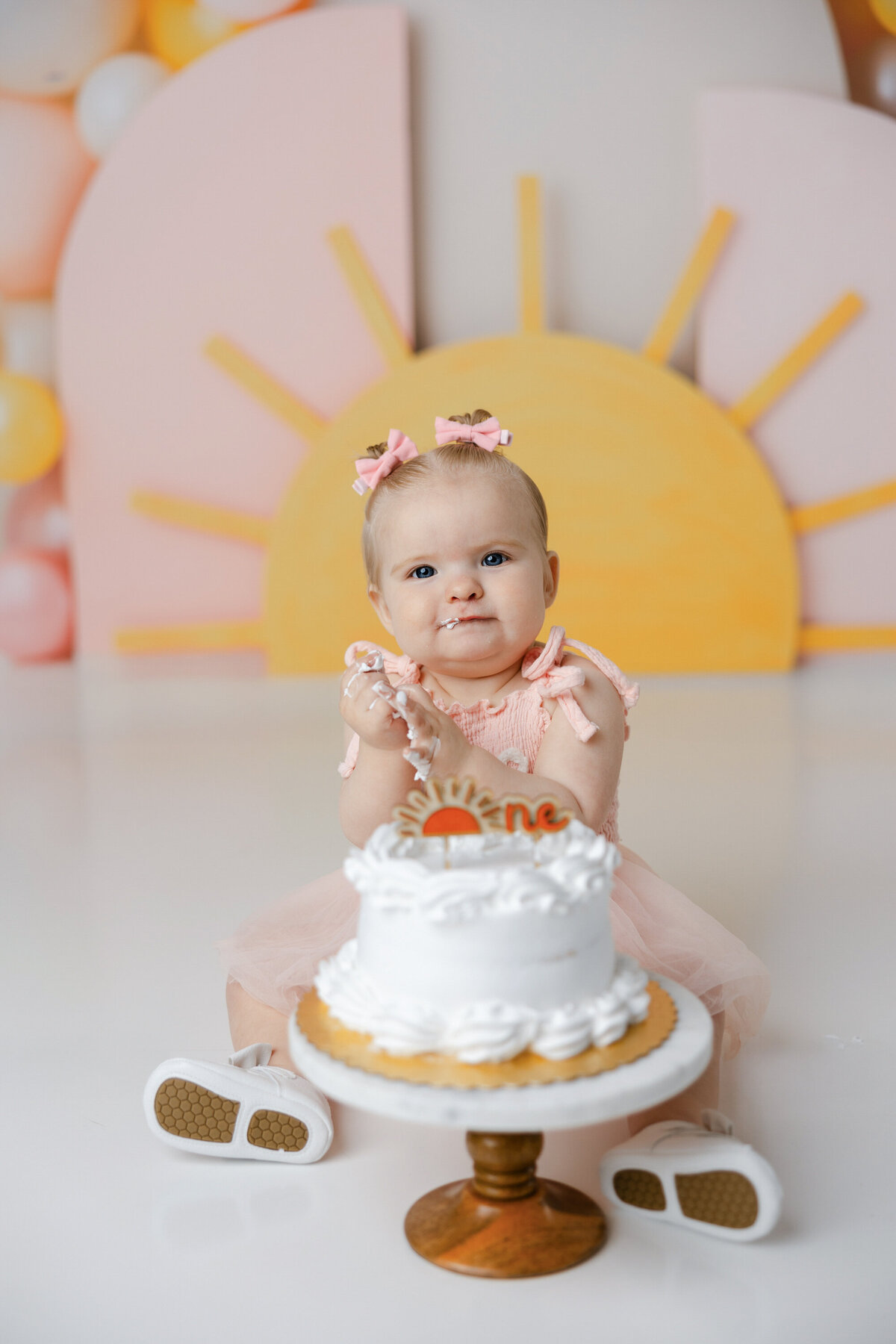 Baby in peach “one” dress sitting with cake during cake smash with sunrise backdrop. Photograph by Yucaipa cake smash photographer Kaitlyn Dawn Photography.