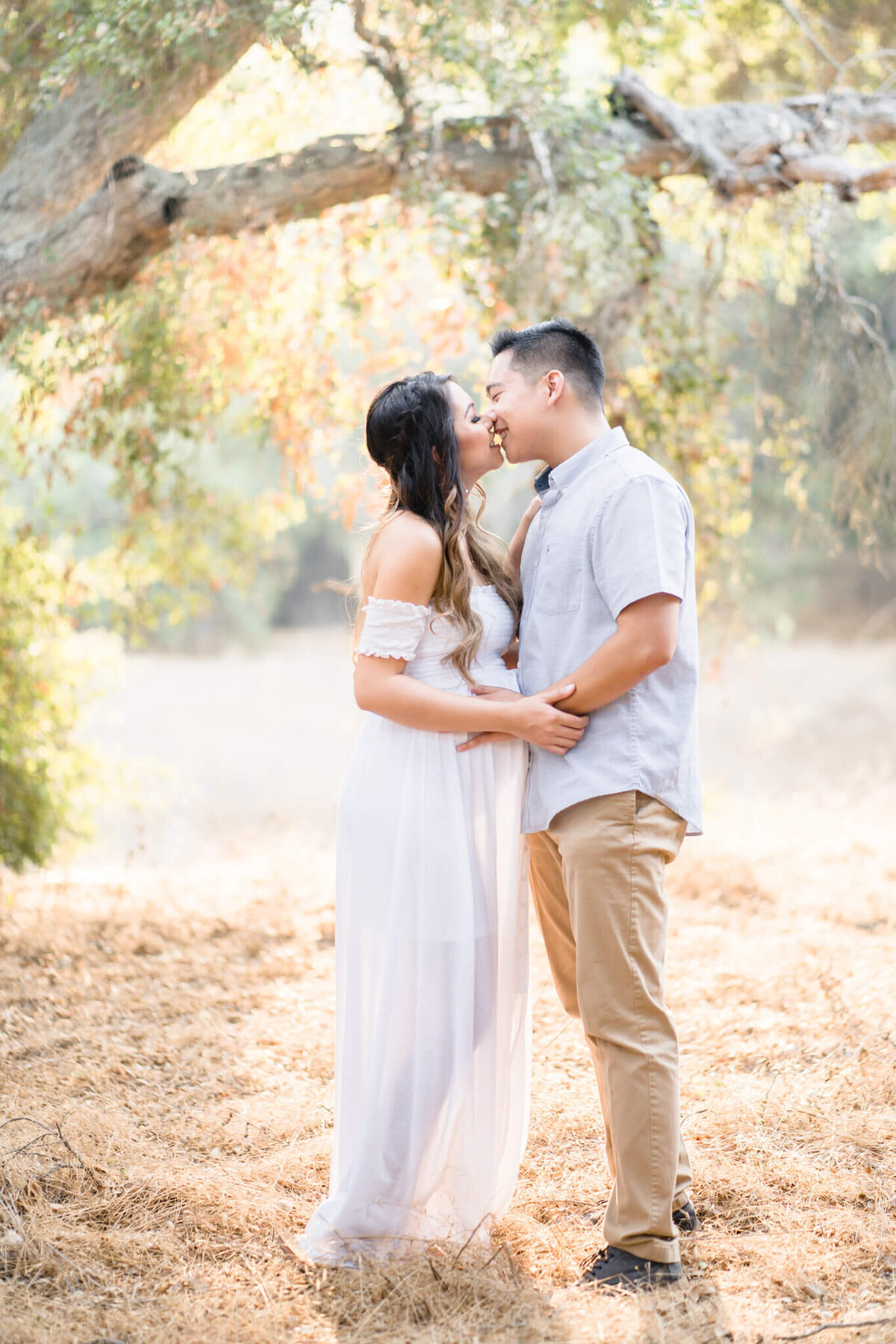 Pregnant mom in white dress holding hands with husband and kissing under autumn tree