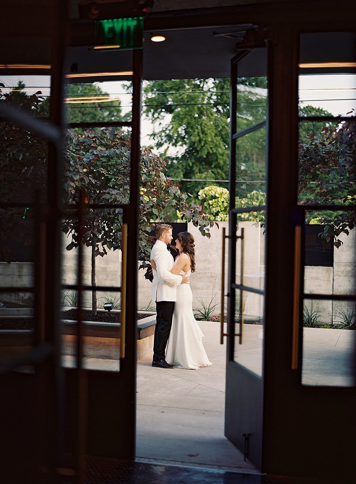 The groom, wearing a tuxedo with a white jacket and the bride wearing a white silk halter top sheath dress embrace in the courtyard of Clementine Hall during their summer wedding as we  look at them through the tall glass doors of the venue.