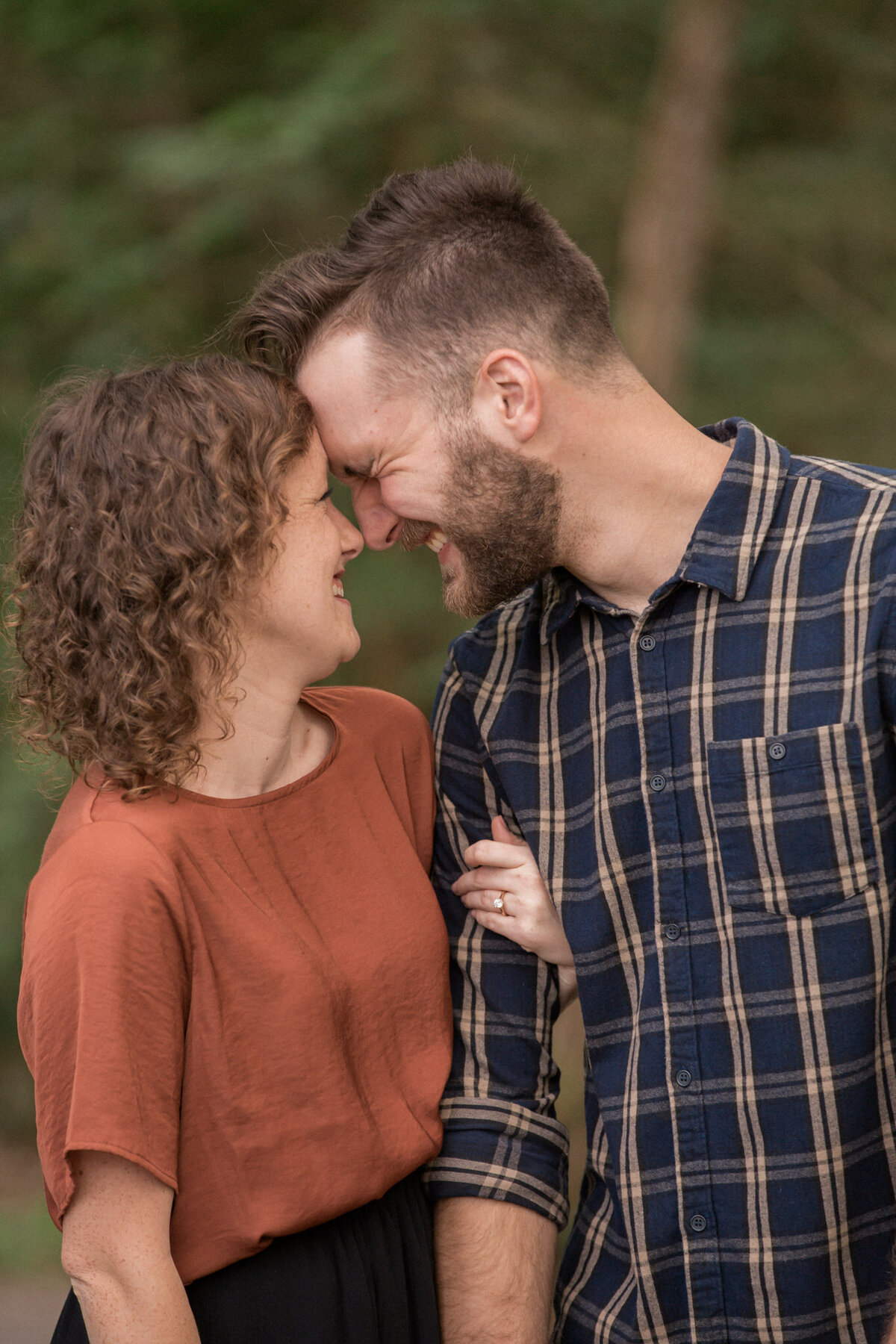 A man wearing blue flannel smiles, squinting his eyes as his fiancee brings her forehead to his.