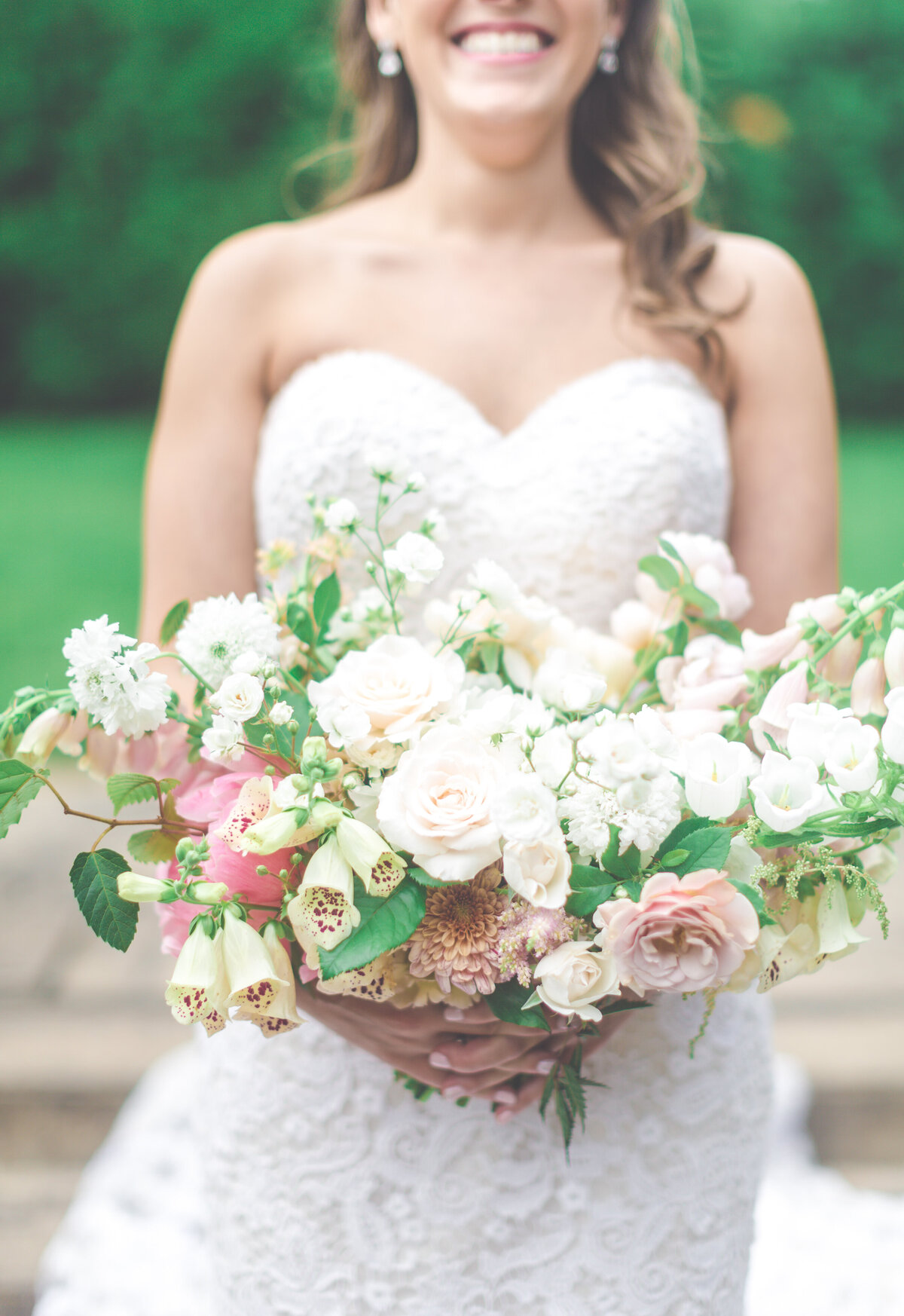 A smiling bride stands in a field holding a lush bouquet of white flowers.