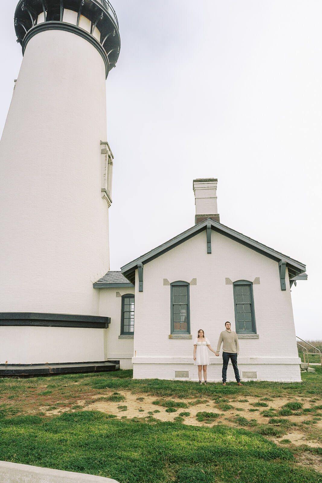 A coule standing and holding hands in front of a foggy lighthouse. 