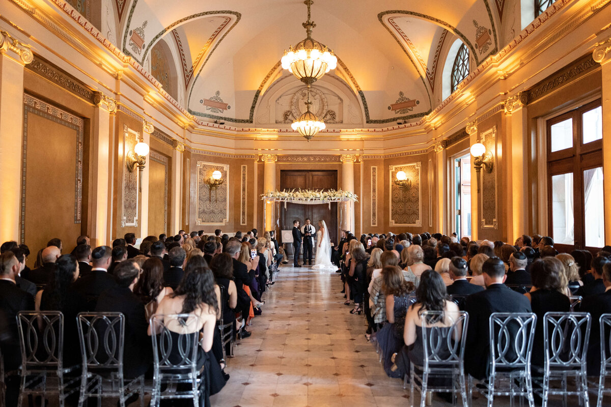 A bride and groom stand at the altar in a grand, elegantly decorated ballroom with guests seated on either side of an aisle. The ceiling features ornate designs and chandeliers, and light streams in through large windows.