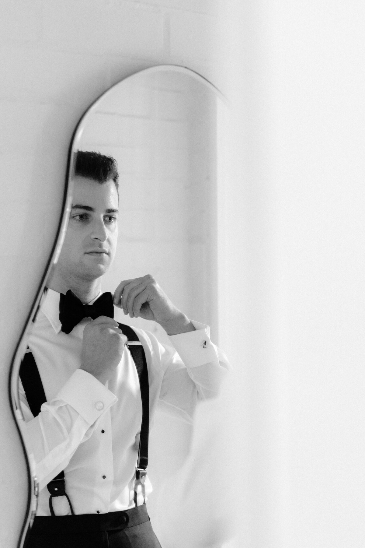 A groom fixing his bow tie in front of a mirror.