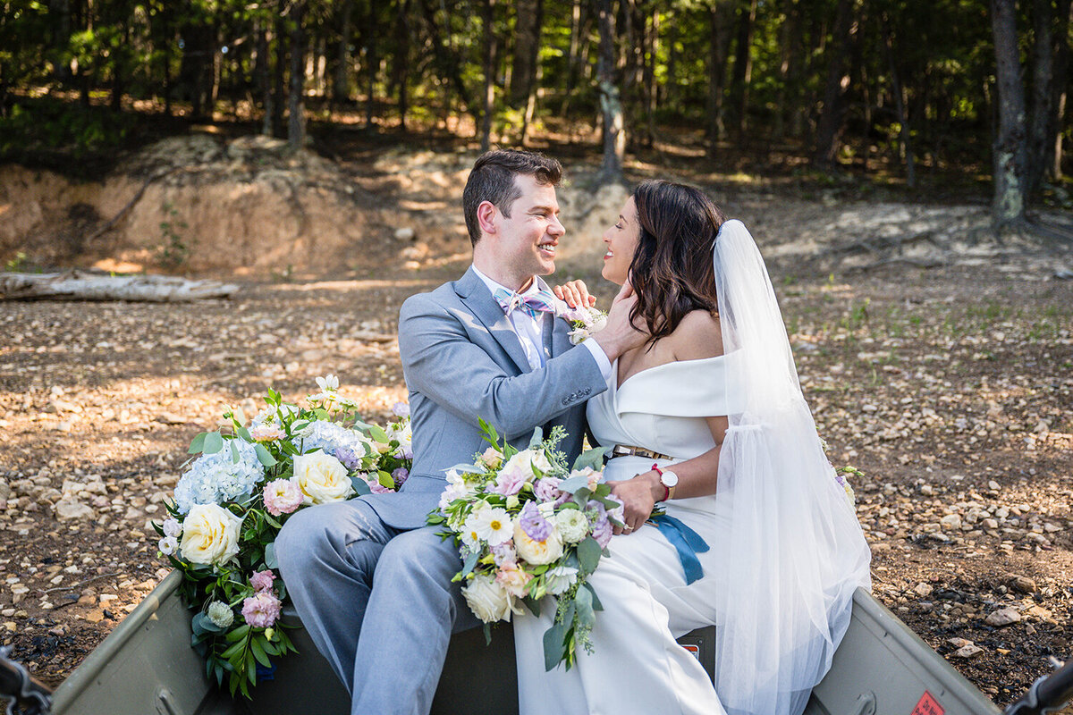A wedding couple sit in a rowboat that is now parked on the shores of Carvin’s Cove in Virginia. The boat is adorned with pastel flowers and the couple sits hip to hip on one of the benches in the boat. The groom places his hand on the bride’s neck while she places a hand on his shoulder. The pair smile widely at one another.