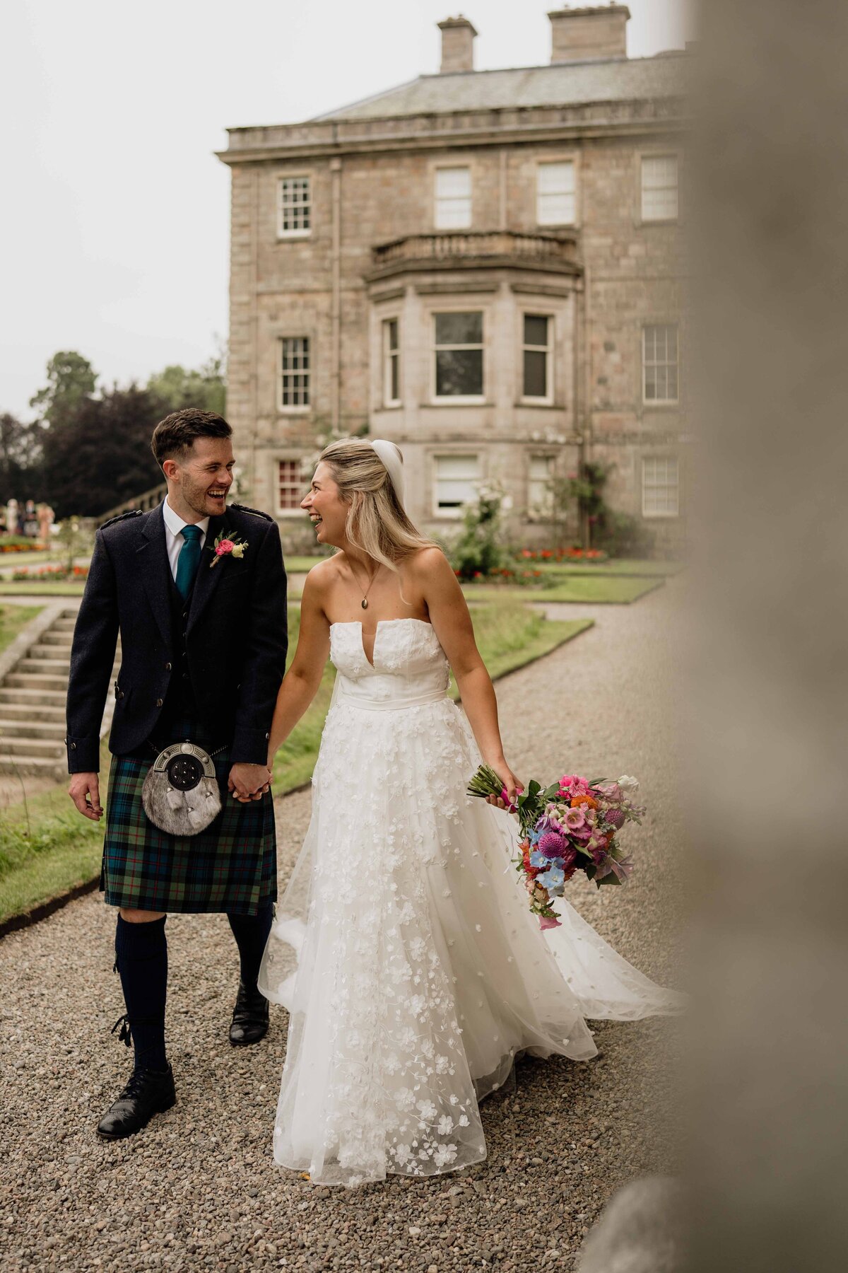 A bride and groom hold hands and walk in front of Haddo House in Aberdeenshire. They laugh with each other.