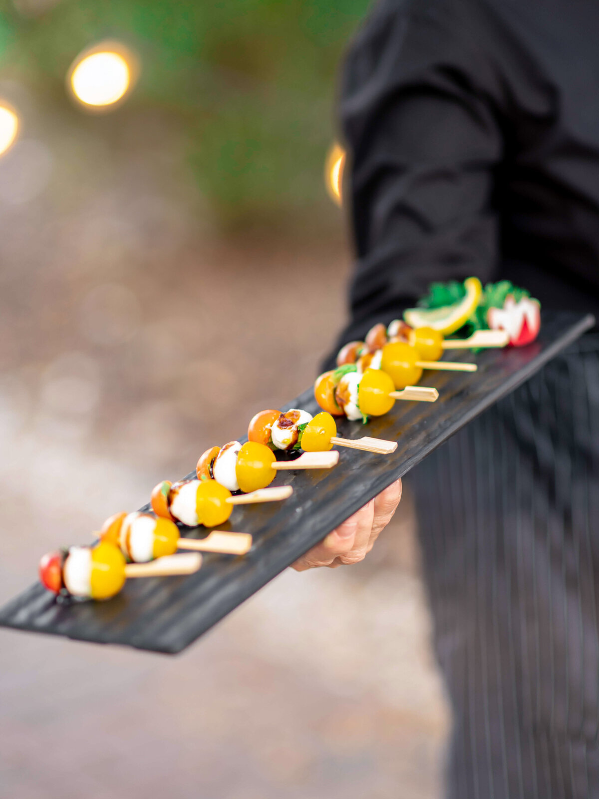 A server in a dark outfit holds a rectangular black tray with various bite-sized appetizers skewered on sticks, including yellow cherry tomatoes. The background is softly blurred with warm lights.
