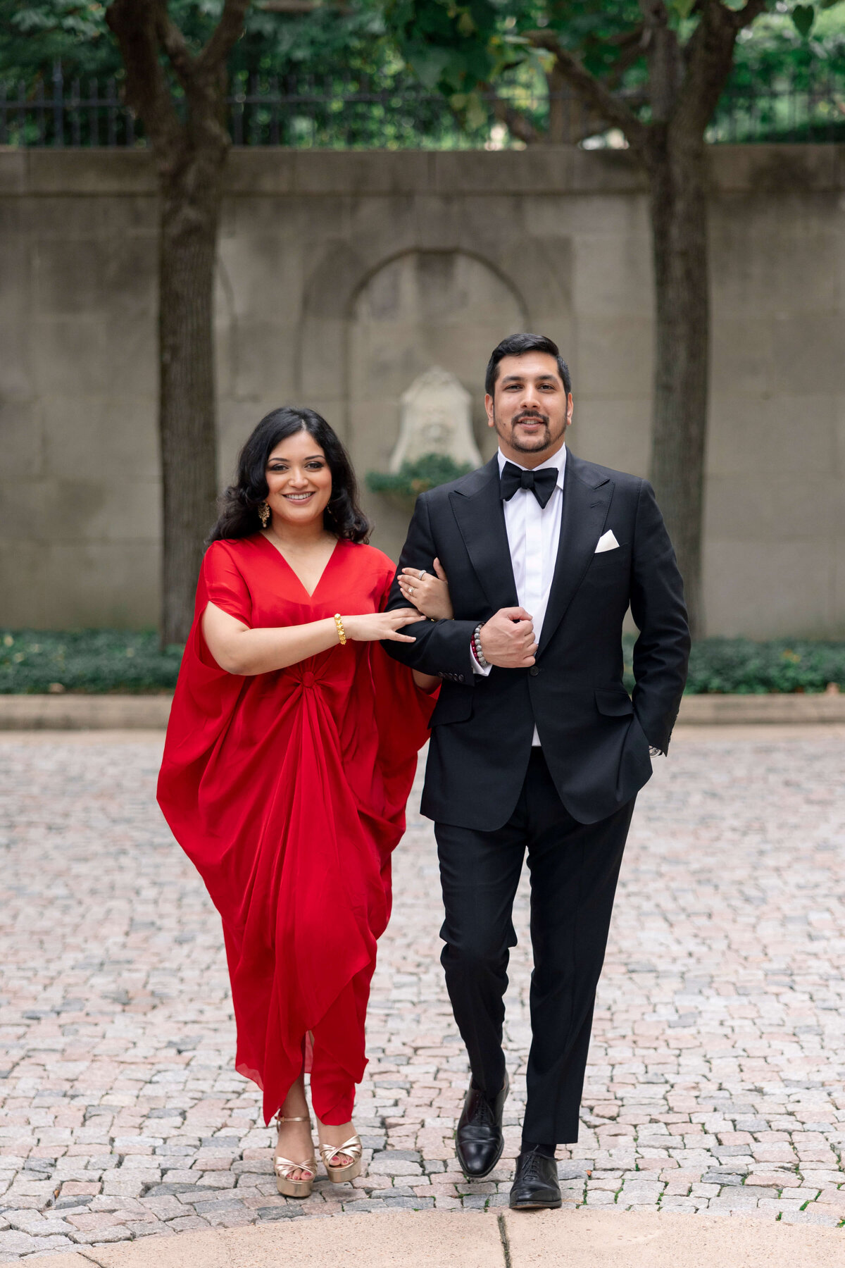 A couple elegantly walks arm in arm outdoors. The woman wears a flowing red dress, and the man is in a black tuxedo with a bow tie. They are smiling and surrounded by a stone walkway and greenery.
