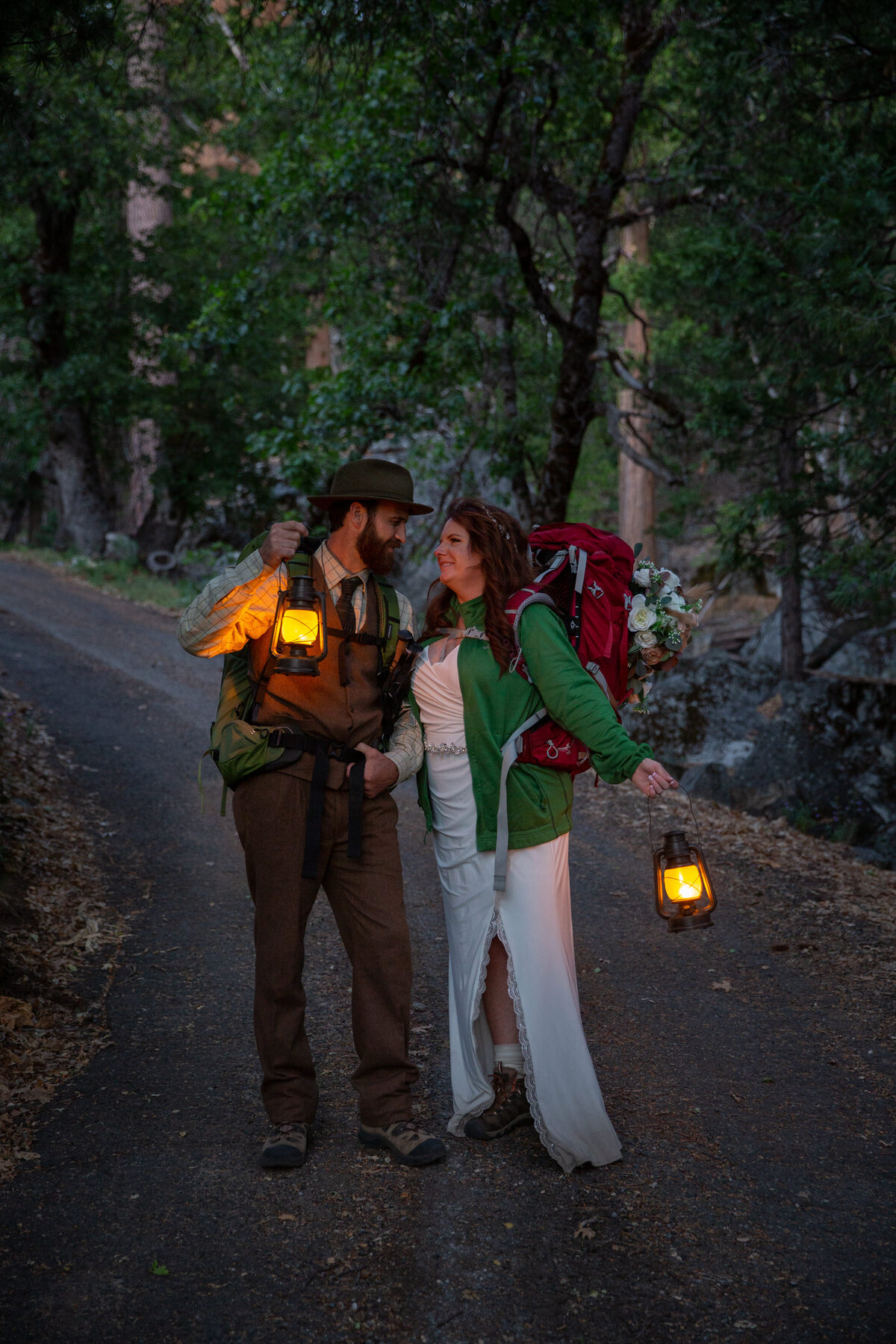 A bride and groom are wearing backpacks and standing on a paved path holding lanterns around them as the sunsets.