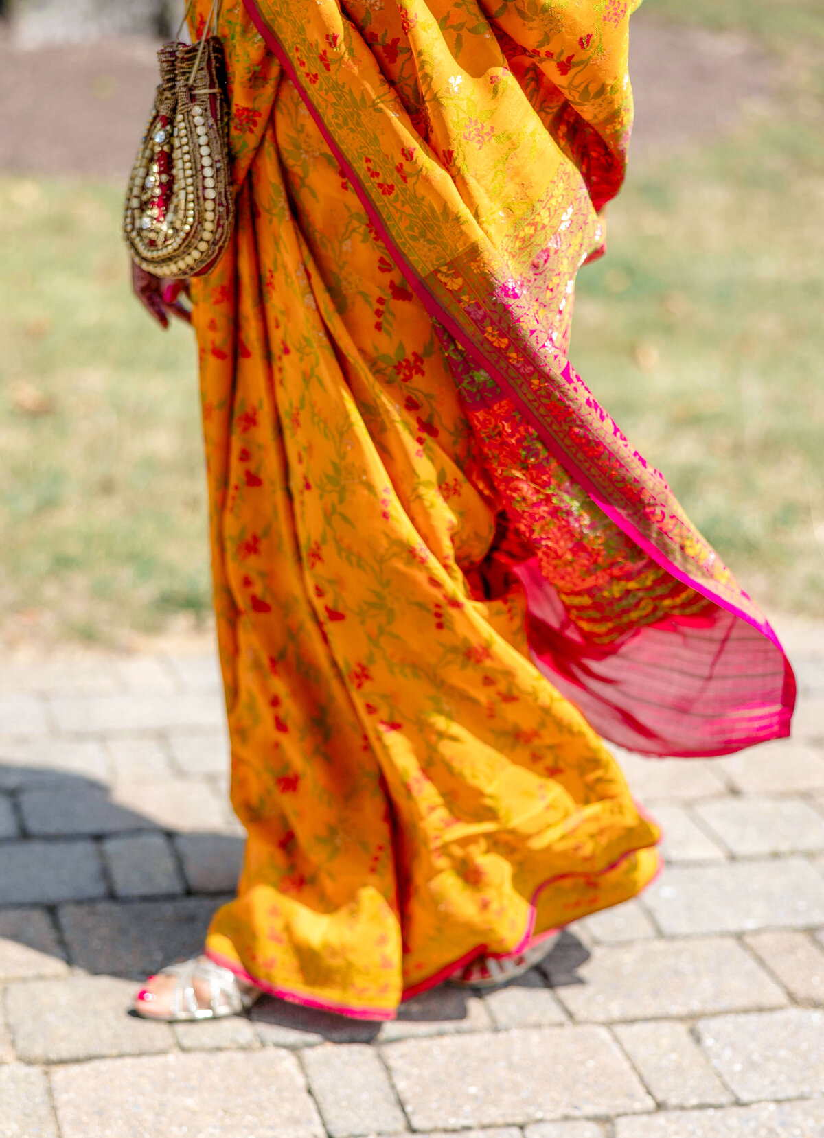 A person wearing a vibrant orange and pink sari walks on a stone pathway. They carry a decorative beaded bag and wear embellished sandals. The sari features a floral design with intricate details.