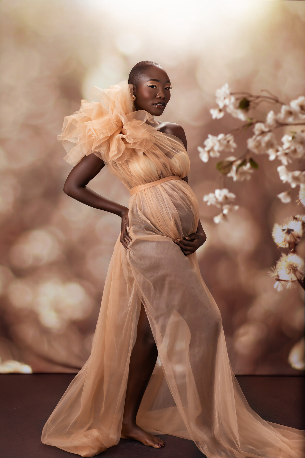 A radiant woman standing gracefully in a flowing sheer golden gown, surrounded by delicate white blossoms. Captured by a DFW maternity photographer, this ethereal portrait exudes elegance and natural beauty.
