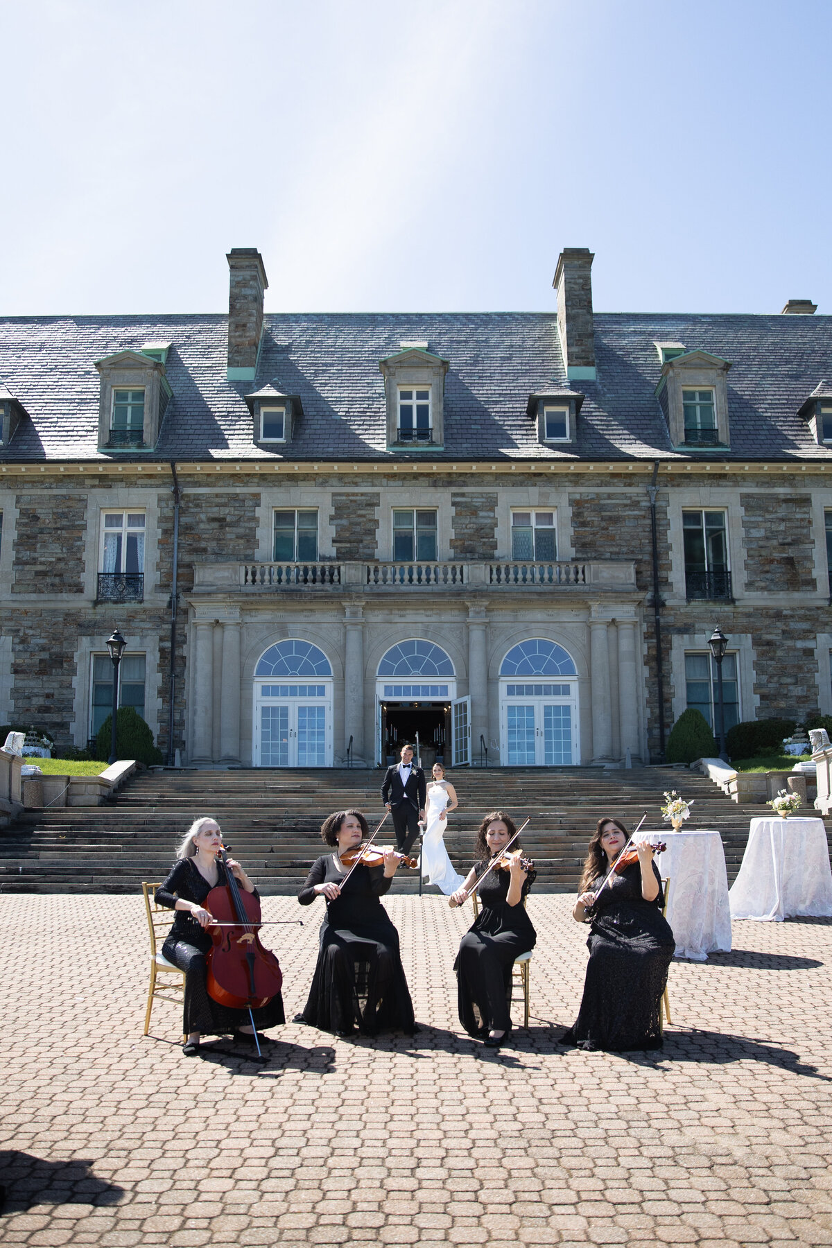 Aldrich Mansion Wedding Photos | Kelsey Sheehan Photography Timeless Rhode Island Weddings | String quartet in black dresses performs at a wedding in front of a grand stone mansion. The bride and groom, smiling, descend steps in the background.