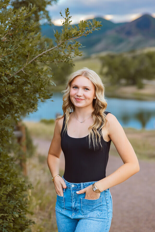 Girl smiles with hands in pockets and a lake behind her.