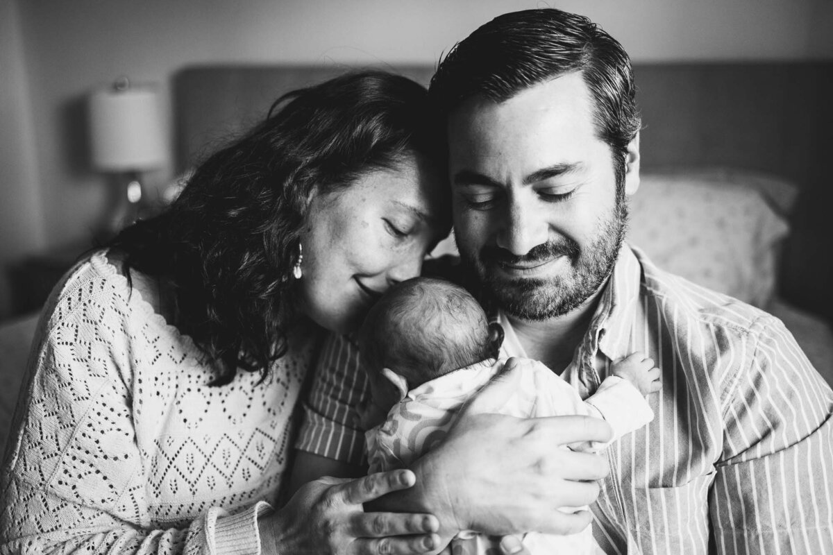 Black & white image of young couple and their newborn daughter sitting at the edge of their bed during a newborn photoshoot.