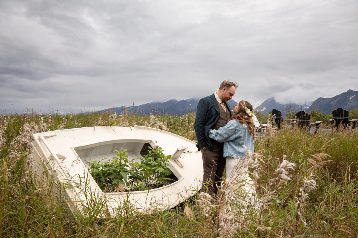 A bride and groom stand in front of a white boat in a field of flowers in Alaska.