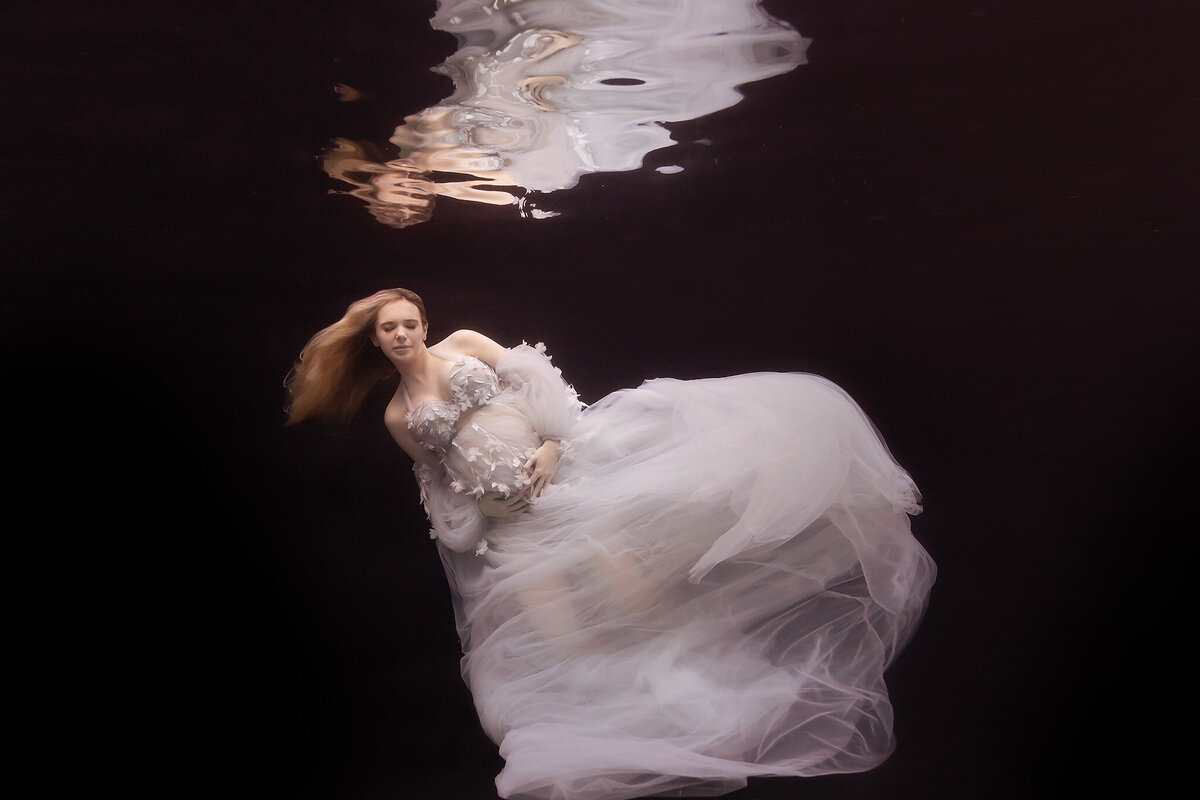 A serene underwater pregnancy photo captured by a Dallas underwater pregnancy photographer. The woman is enveloped in a voluminous white gown adorned with delicate floral details, her hair floating gracefully as she cradles her belly. The soft, flowing fabric creates a dreamlike effect against the dark backdrop.