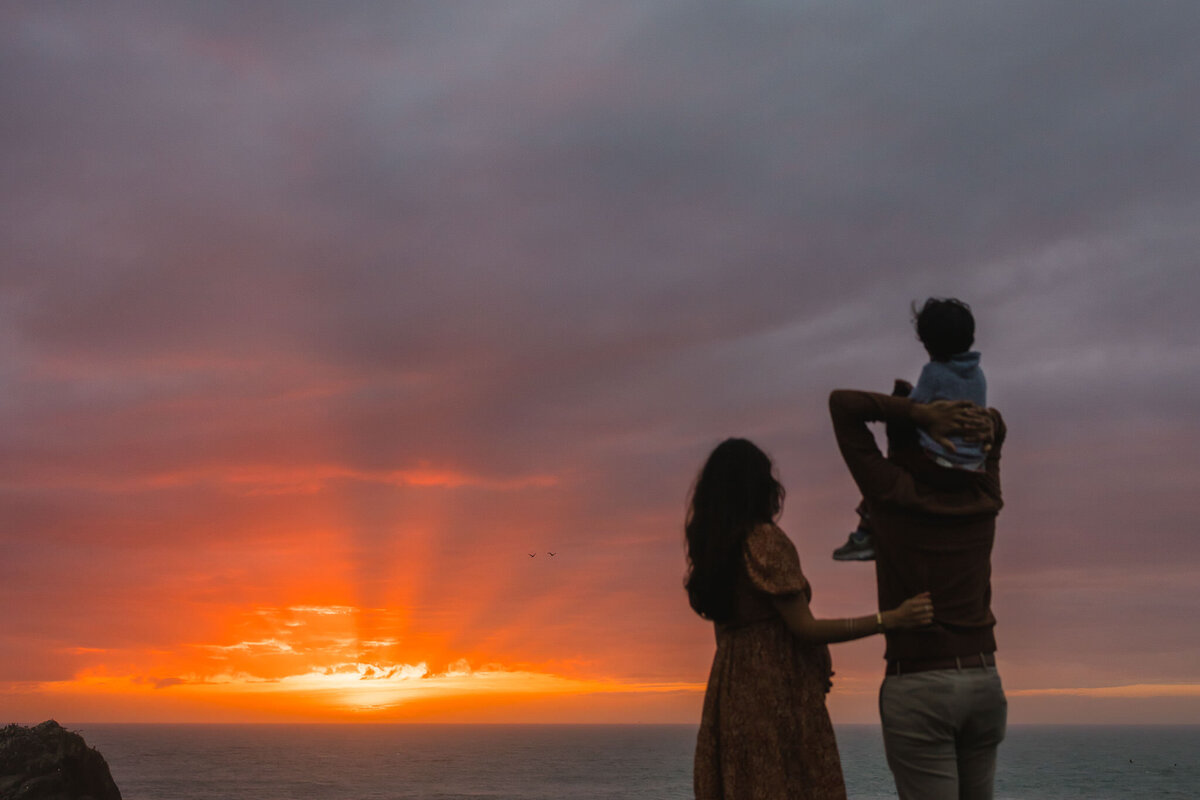 Documentary family photo at sunset. Family looks toward ocean as sun beams break through a cloudy sky