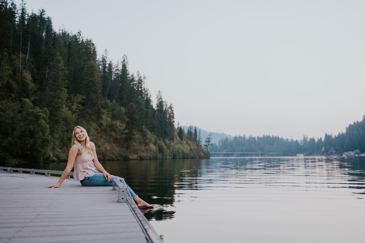 Young woman smiles while sitting on dock and swinging feet into Coeur d'Alene lake.