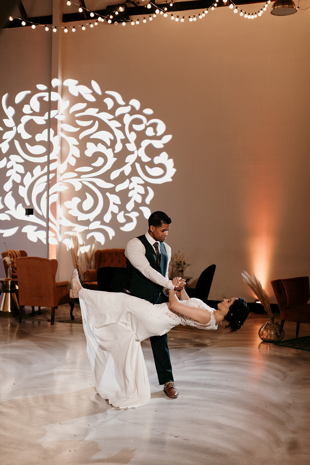 Groom dipping the bride on the dance floor, with a projected floral design on the wall and string lights above, adding a magical touch.