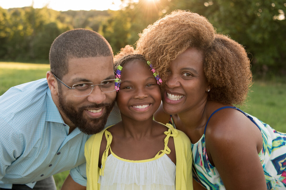 Two parents are pressing their cheeks against their young daughter's face and smiling directly at the camera with the sun setting in the field behind them