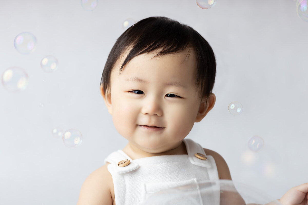 A joyful baby, dressed in a white romper, sits amidst floating bubbles during a milestone session with a Dallas milestone photographer, captured against a soft, neutral backdrop.