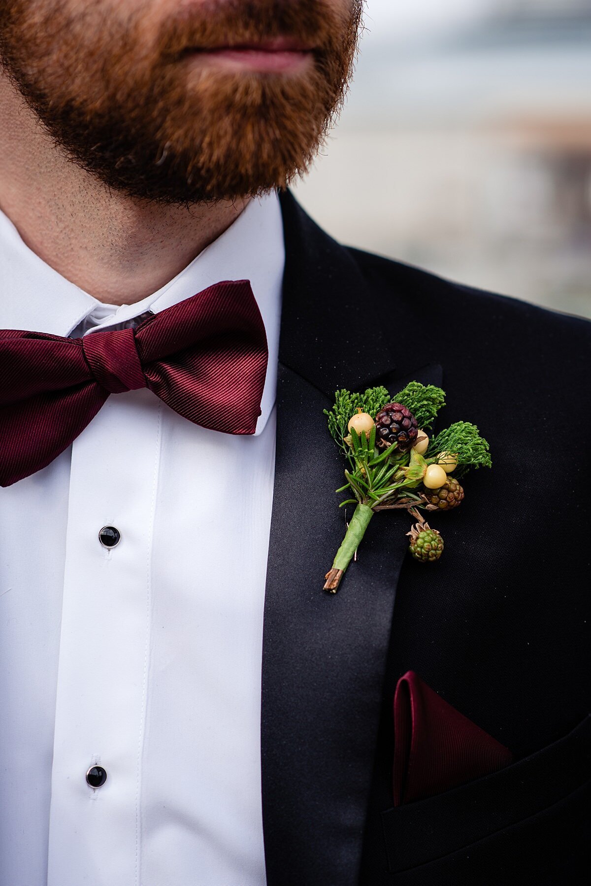 A groom wearing a black tuxedo with a white shirt with black buttons and a burgundy bowtie has a boutonniere made from fresh fruit and vegetables. The boutonniere is made from broccoli, white berries and black raspberries.