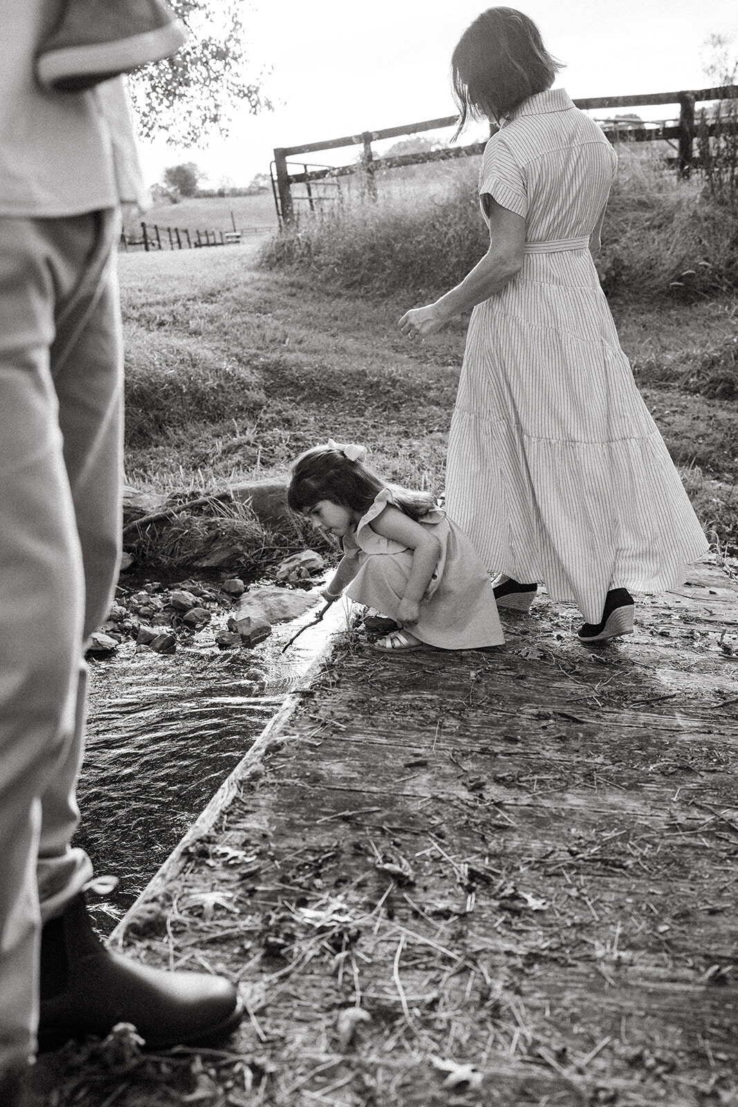 Toddler girl splashing in a shallow creek while mom stands behind her during a documentary-style session
