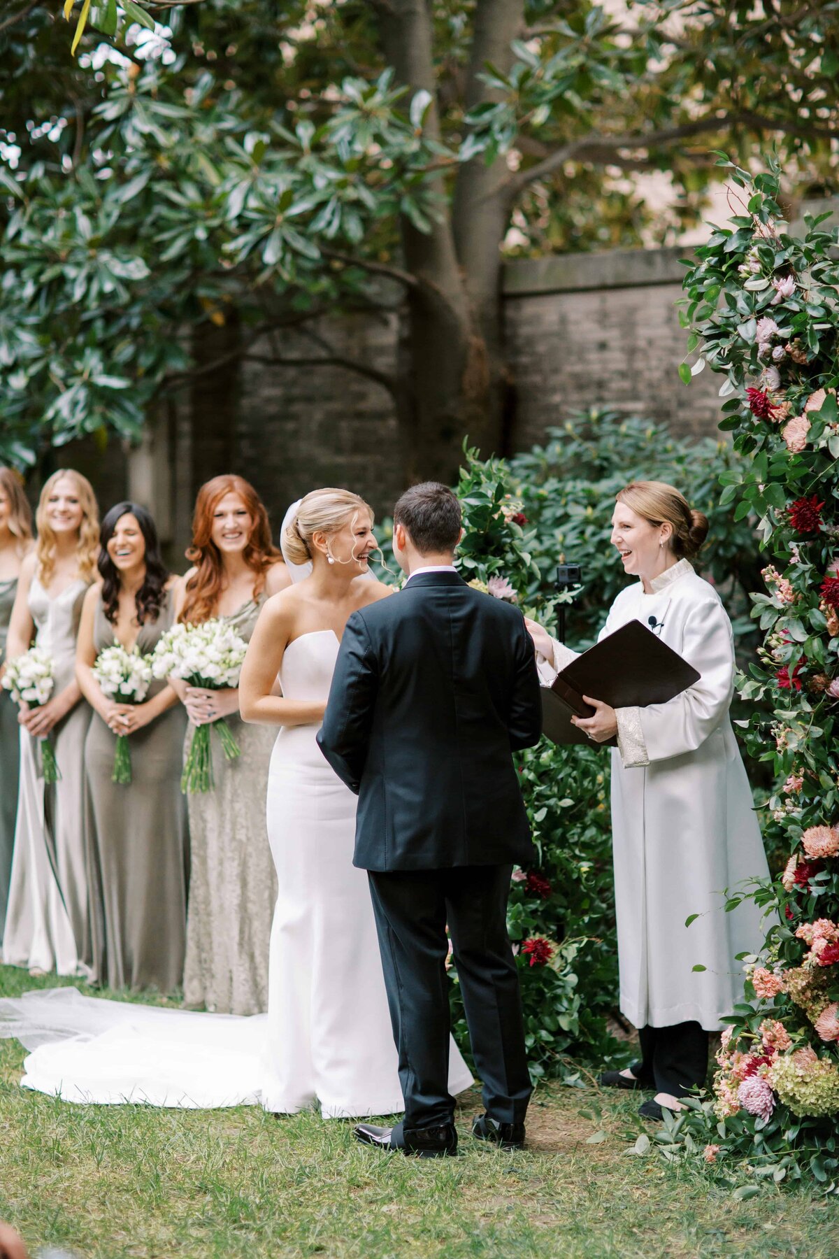 A fair haired bride is laughing while holding the hands of a handsome groom in a black tuxedo as they get married with the officiant beside them at the Larz Anderson House.
